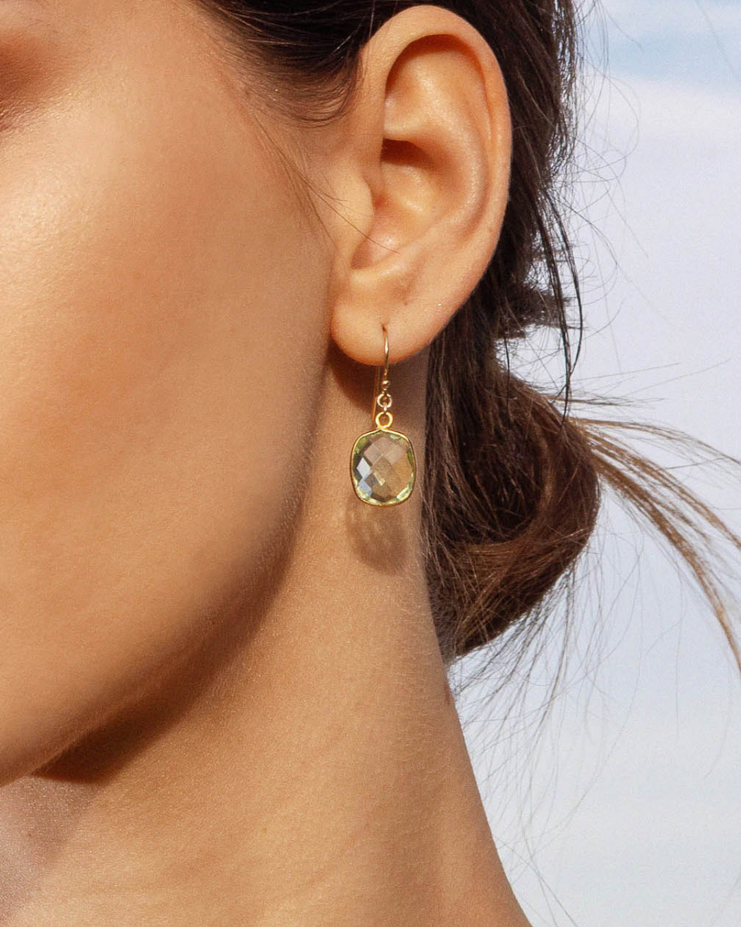 Close-up of a woman's ear wearing a gold earring with a green amethyst bezel gemstone against a blurred background.