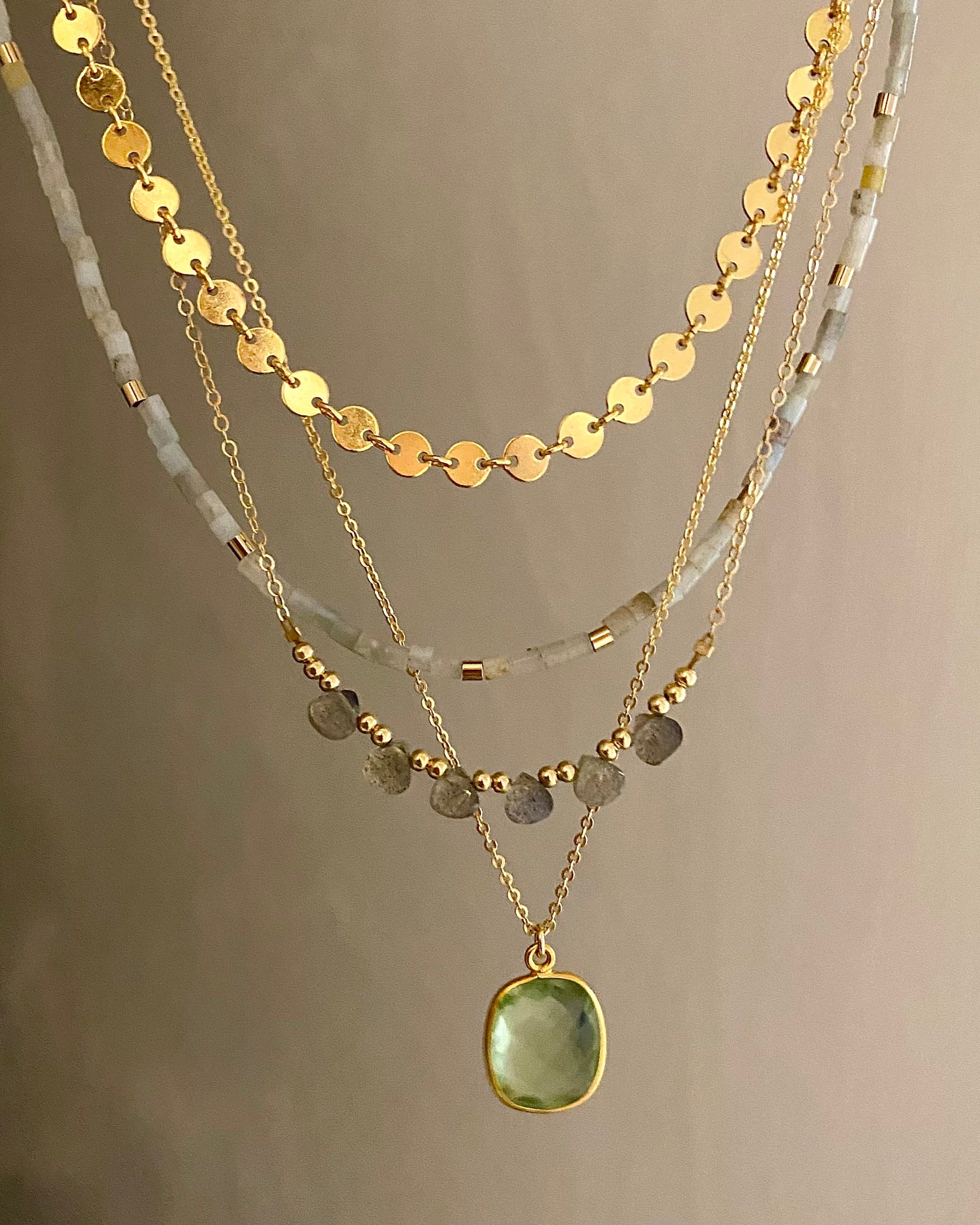 Gold gemstone necklaces with one featuring a green amethyst pendant on a neutral background