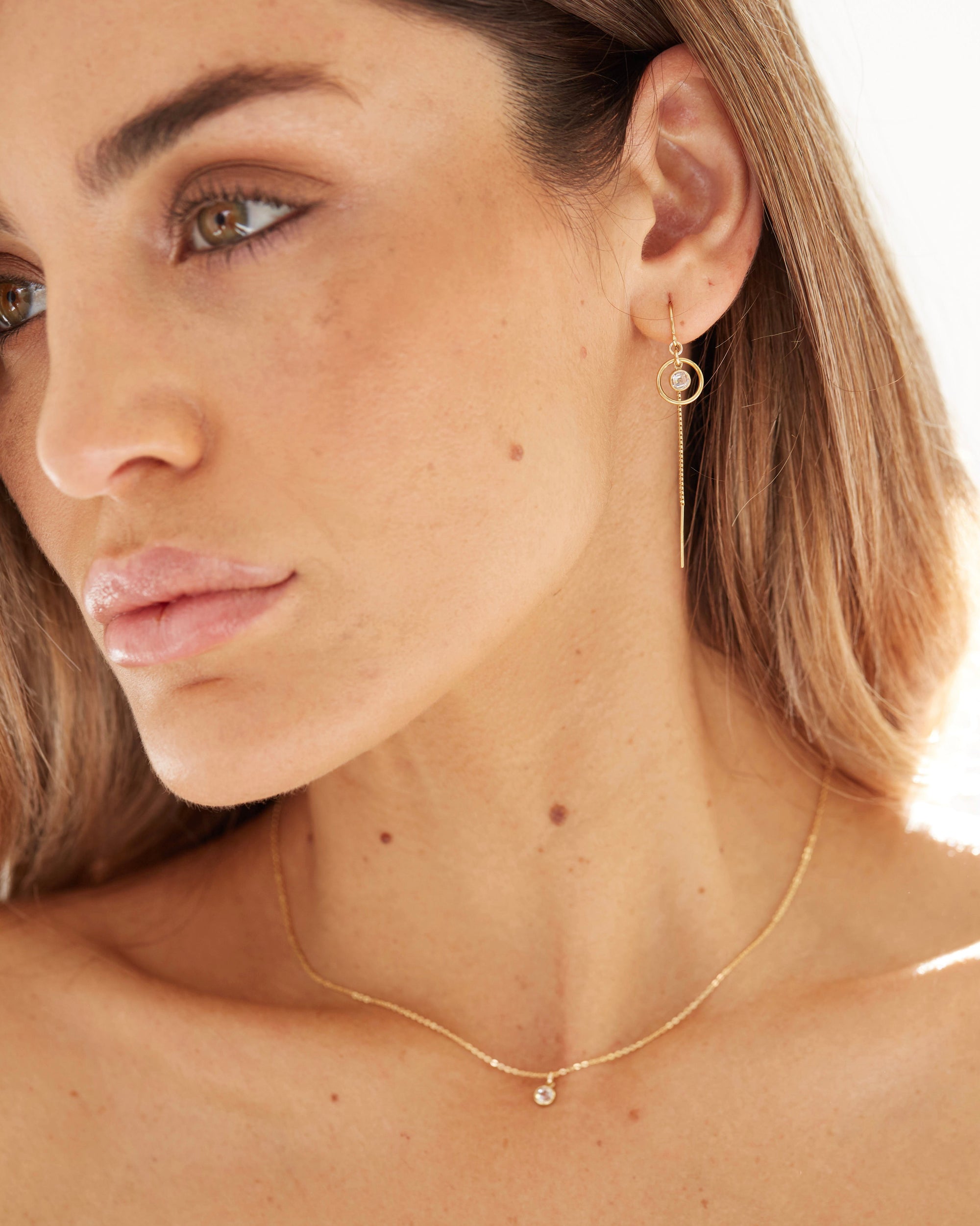 Close-up of a woman wearing gold thead cubic zirconia earrings and delicate necklace against a neutral background