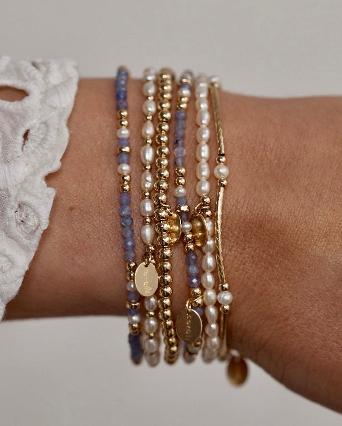 Close-up of a wrist wearing multiple pearl and gemstone gold beaded bracelets with a neutral background