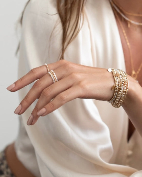 Close-up of a woman wearing a cream satin top with gold and pearl bracelets and rings with a neutral background